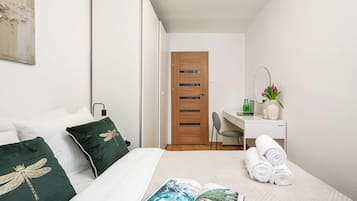 A hotel room with a bed, a desk, a chair, a wooden door, and a small table with a vase of flowers.