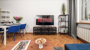 Een woonkamer met een televisie waarop 'NETFLIX' staat, een eethoek met stoelen en een tafel, en een boekenkast met boeken.
