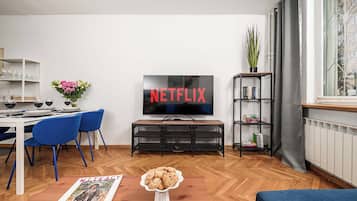 Een woonkamer met een televisie waarop 'NETFLIX' staat, een eethoek met stoelen en een tafel, en een boekenkast met boeken.