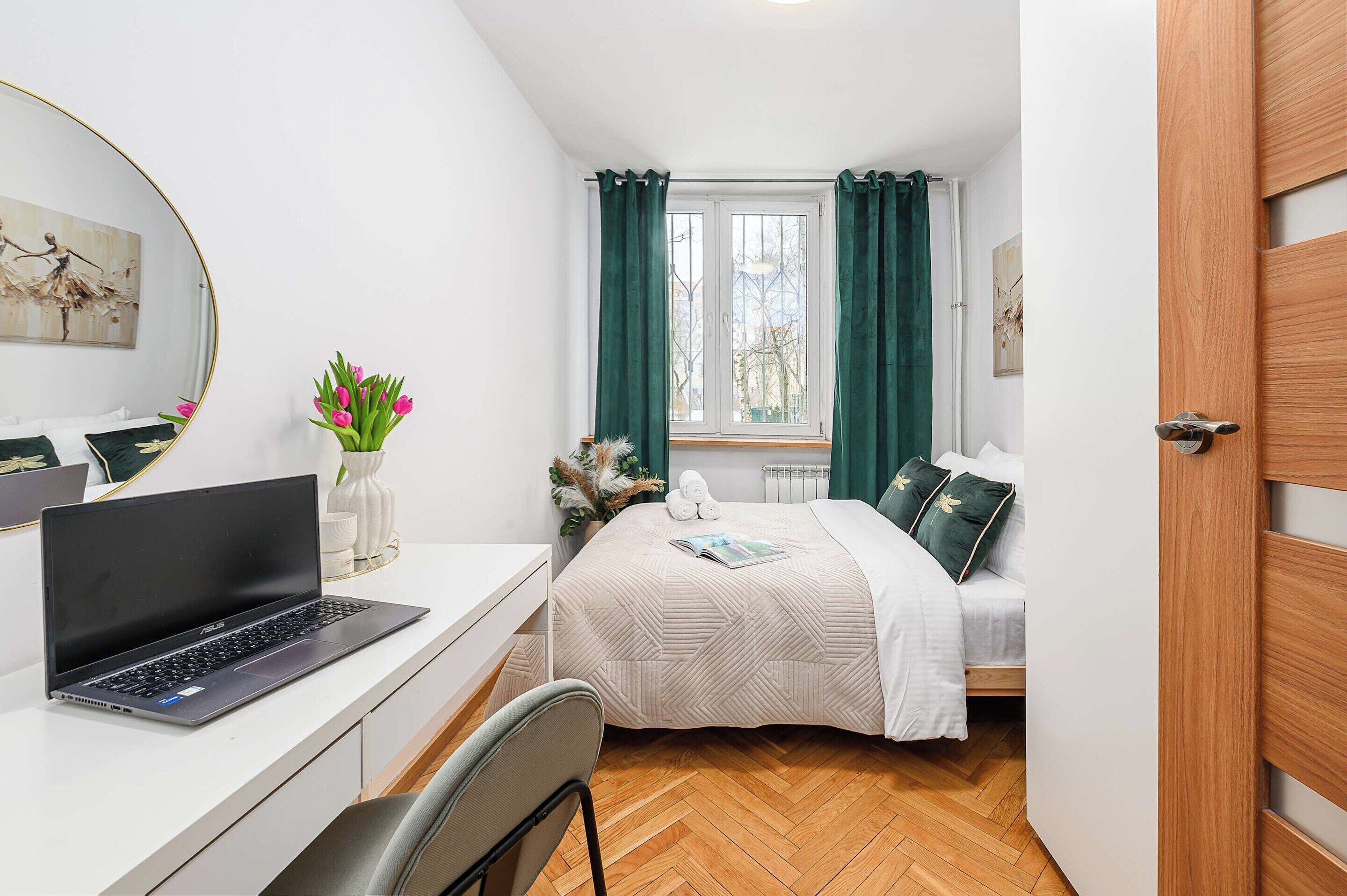Une chambre à coucher avec un lit, un bureau avec un ordinateur portable, une chaise, une fenêtre avec des rideaux et une porte.