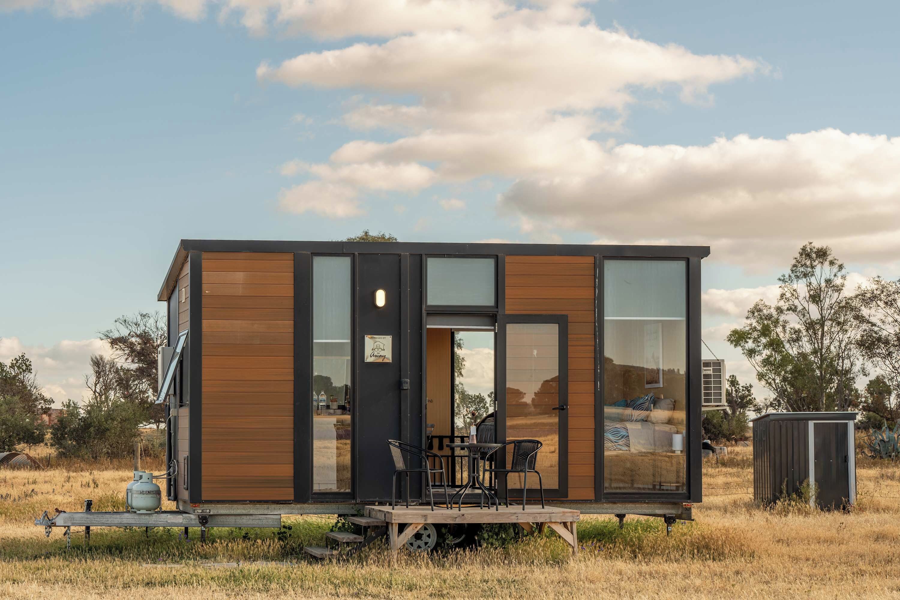 Una cabaña moderna y compacta con fachada de madera, ventanas amplias y una pequeña terraza con mobiliario exterior.