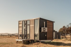 A stationary RV with a sliding door, a small porch with chairs, and an air conditioning unit mounted on the side. - Alpaca Paddock X by Tiny Away (Peterborough)