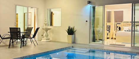 A modern indoor swimming pool with a clear view of the adjacent room, a ceiling fan, and a dining area with chairs and a table.