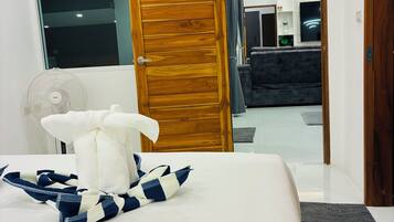 A hotel room with a bed, a wooden door, a fan, and a view into the living area.