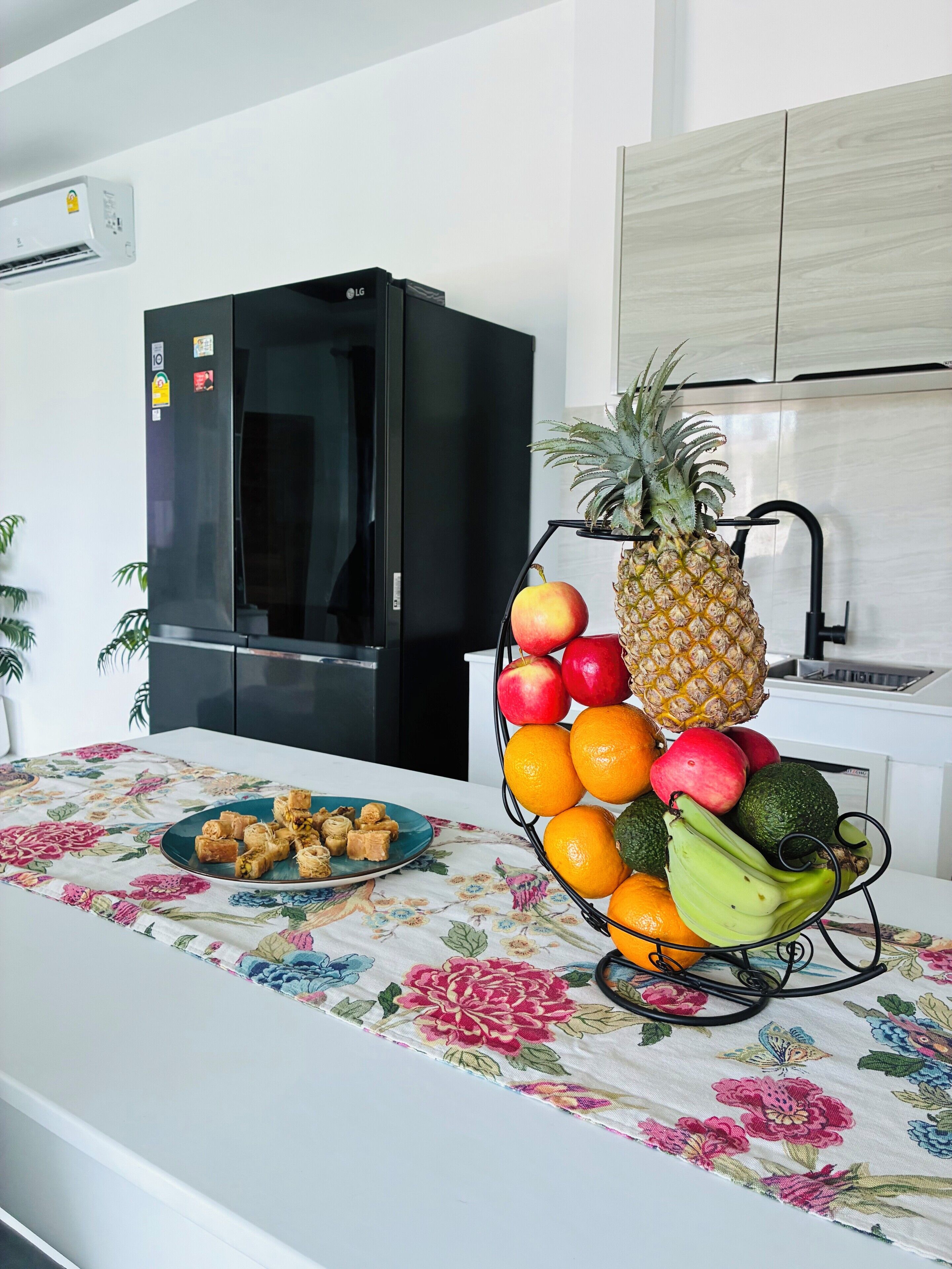 Una cocina con un refrigerador negro, una cesta de fruta en la encimera y un mantel con motivos florales.
