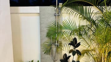 A small outdoor shower area with a metal showerhead, two potted plants, and a concrete wall.