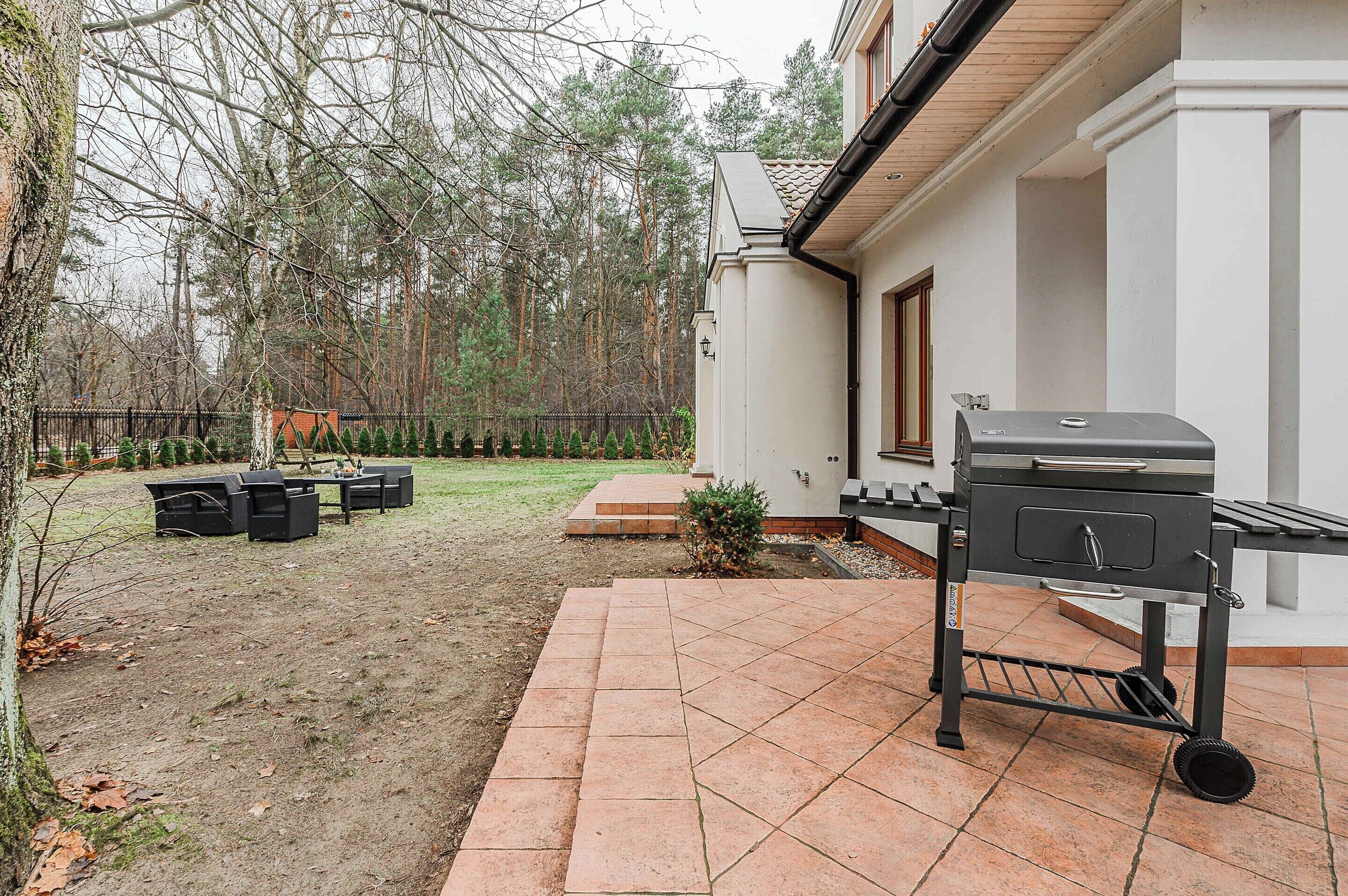 Un patio con una parrilla, mobiliario exterior y un pequeño jardín con árboles y una cerca.