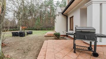 Un patio con una parrilla, mobiliario exterior y un pequeño jardín con árboles y una cerca.