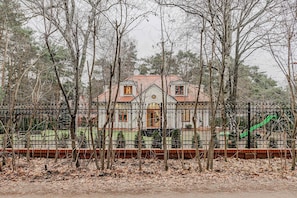 Una casa con techo de tejas rojas, rodeada por una cerca de metal negro, y un tobogán verde al fondo.