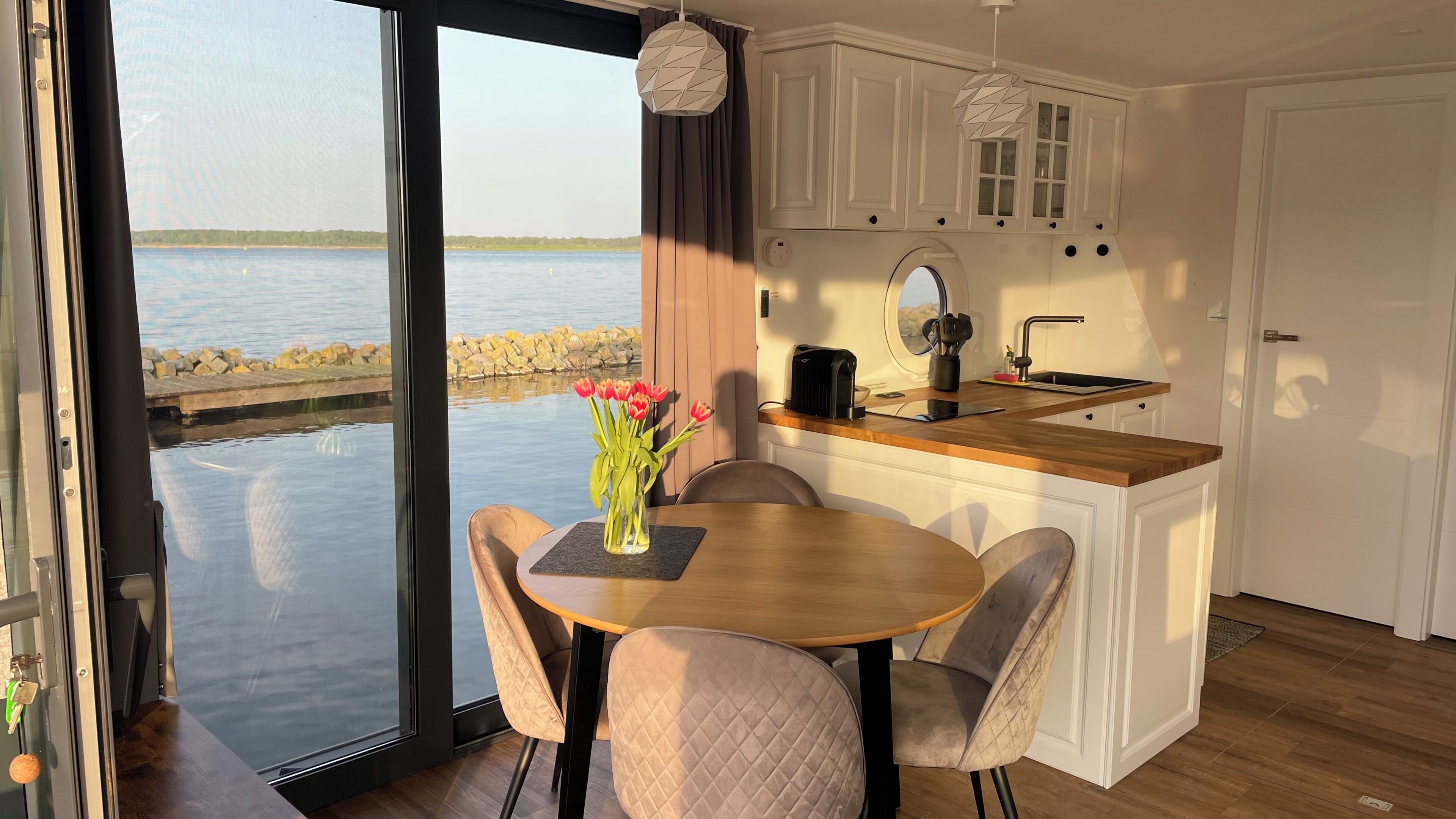 Eine moderne Küche mit einer Holzausziehtheke, einem Spülbecken und Schränken. Ein Essbereich mit Tisch und Stühlen lädt zum Verweilen ein, dazu steht eine Blumenvase. Durch das Fenster ist ein Blick auf das Wasser möglich.