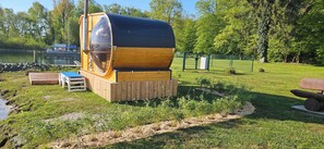 A large cylindrical structure with a reflective surface, surrounded by a grassy area with a wooden bench and a small table. - Modernes festliegendes Hausboot mit großzügiger Dachterrasse und Ruderboot (Röbel/Müritz)