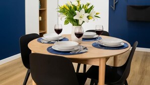 Comedor con una mesa redonda dispuesta para cuatro personas, rodeada de sillas negras. Hay dos platos blancos, dos copas de vino y un jarrón con flores blancas sobre la mesa.