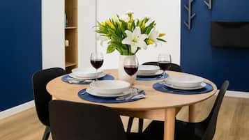 A dining area with a round table set for four, surrounded by black chairs. There are two white plates, two wine glasses, and a vase with white flowers on the table.