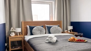 Una cama bien tendida con un edredón gris, dos lámparas de noche, una mesita de madera con un florero y una lámpara, y una bandeja con croissants y fresas sobre la cama.