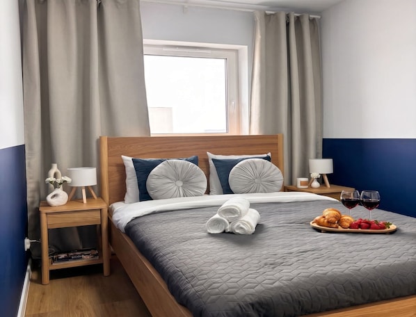 Una cama bien tendida con un edredón gris, dos lámparas de noche, una mesita de madera con un florero y una lámpara, y una bandeja con croissants y fresas sobre la cama.