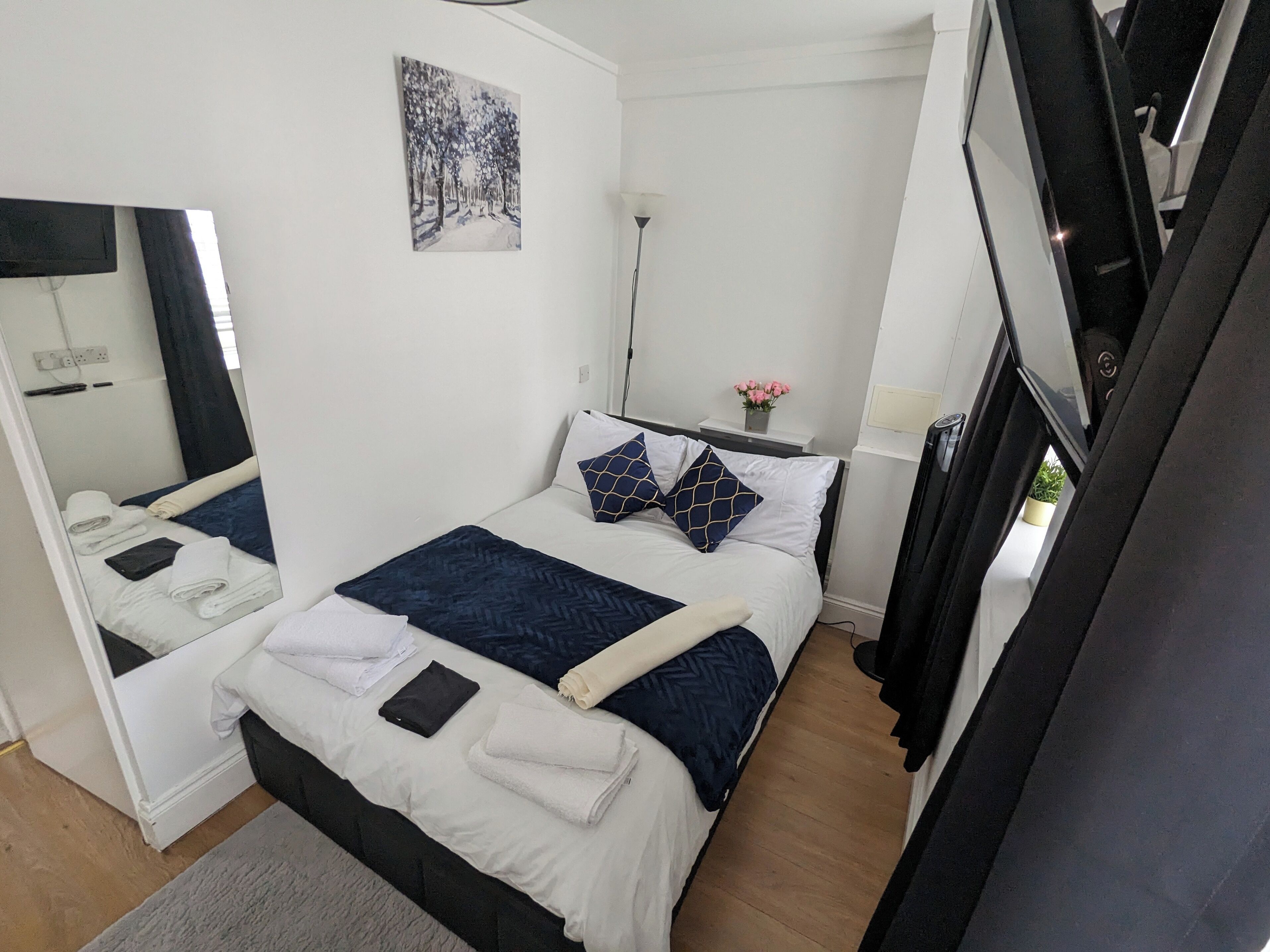 A hotel room with a bed, a mirror, a painting, a bedside table with a lamp, and a potted plant.