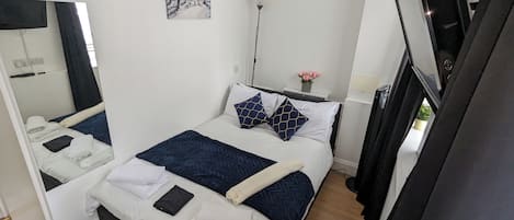 A hotel room with a bed, a mirror, a painting, a bedside table with a lamp, and a potted plant.