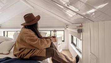 Une personne assise sur un lit, dans une pièce avec une fenêtre, tenant un verre.