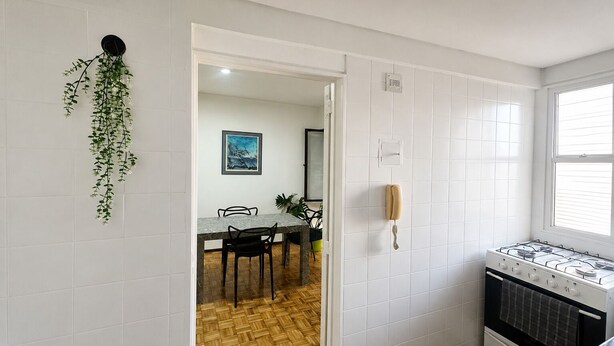 Una cocina con paredes de azulejos blancos, piso de madera, estufa a gas y un comedor con sillas y mesa.