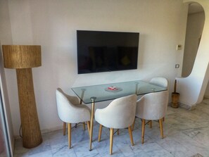 Ein Essbereich mit einem Glastisch, vier Stühlen, einem an der Wand befestigten Fernseher und einer stehenden Stehlampe.