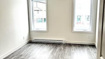 Une pièce avec deux fenêtres, une bouche d’aération visible et un plancher en bois franc.