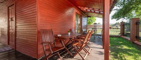 Eine überdachte Terrasse im Freien mit einem Holztisch und Stühlen, einer Tür und Blick auf einen umzäunten Bereich.