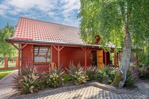 A red wooden house with a tiled roof, surrounded by a garden with various plants and a paved walkway. - Two-story Holiday Home With a Terrace (Darlowo)