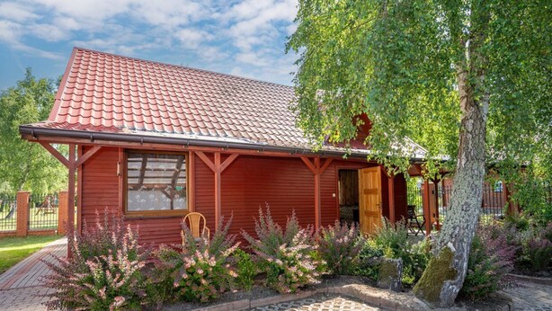 Ett rött trähus med ett tak täckt av tegelpannor, en inglasad veranda och en trädgård med blommande växter.