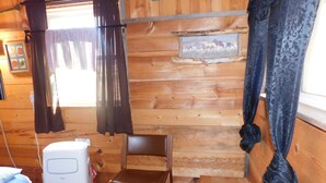 Ein Holzblockhaus-Interieur mit einem Bett, einem Sessel, einem Heizgerät und einem Fenster mit Vorhängen.