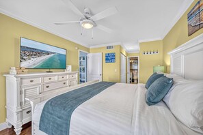 Une chambre à coucher comprenant un lit, une commode, un téléviseur, un ventilateur de plafond et une porte donnant sur une salle de bain.