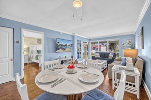 A dining area with a round table set for four, a living room with a sofa and a TV, and a hallway leading to a bedroom. - Dans Florida Condos Beach Beckons at Runaway Bay (Bradenton Beach)