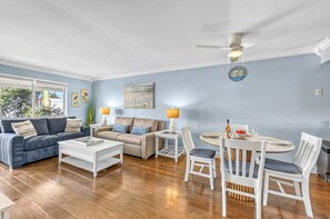 Un salon comprenant un canapé, une table basse, une table à manger avec des chaises, un ventilateur de plafond et des œuvres d’art accrochées aux murs.