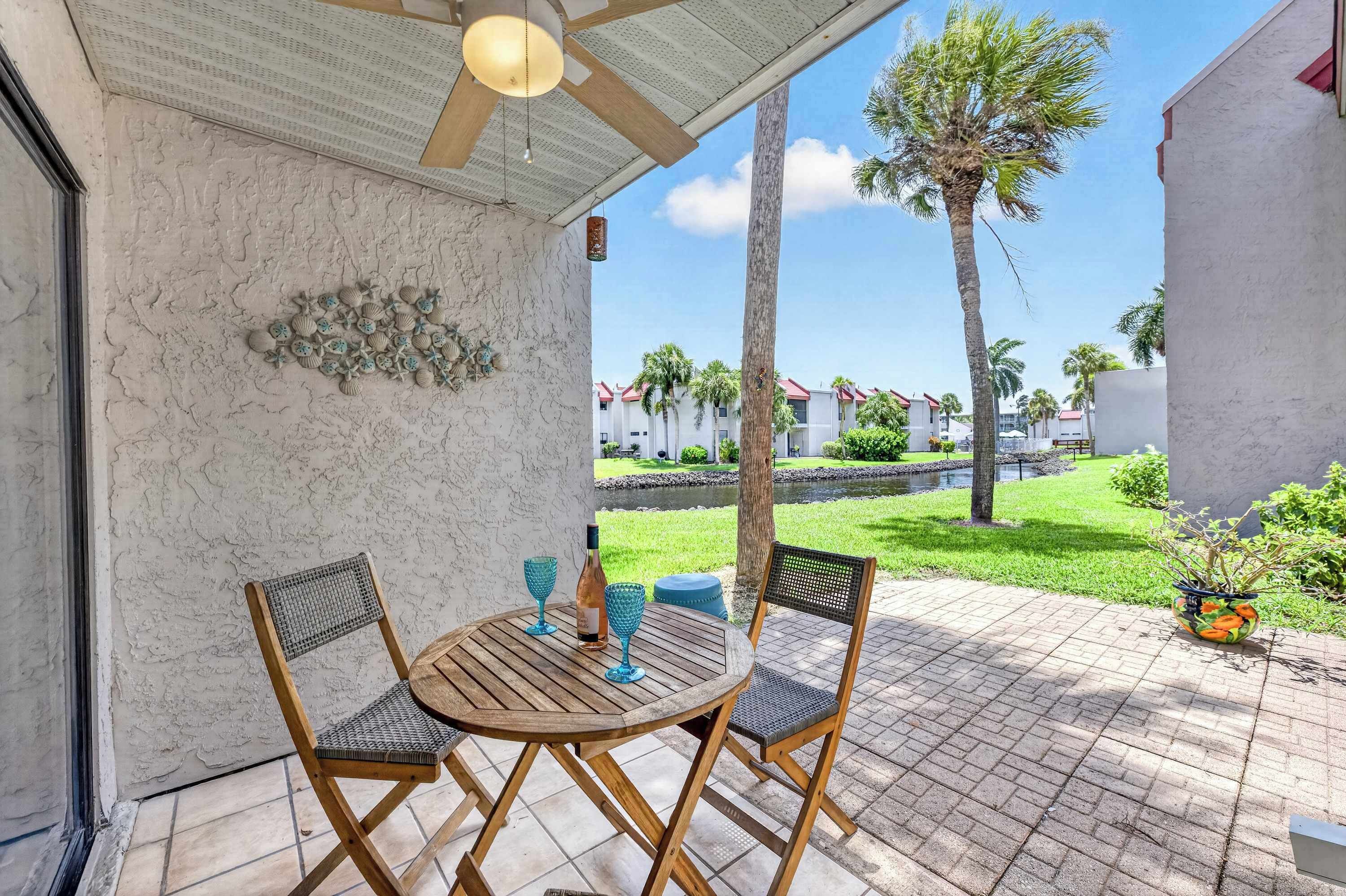 Eine Terrasse mit Holztisch und Stühlen, eine Zierwand mit Muscheln und ein Blick auf einen Pool und Gebäude im Hintergrund.
