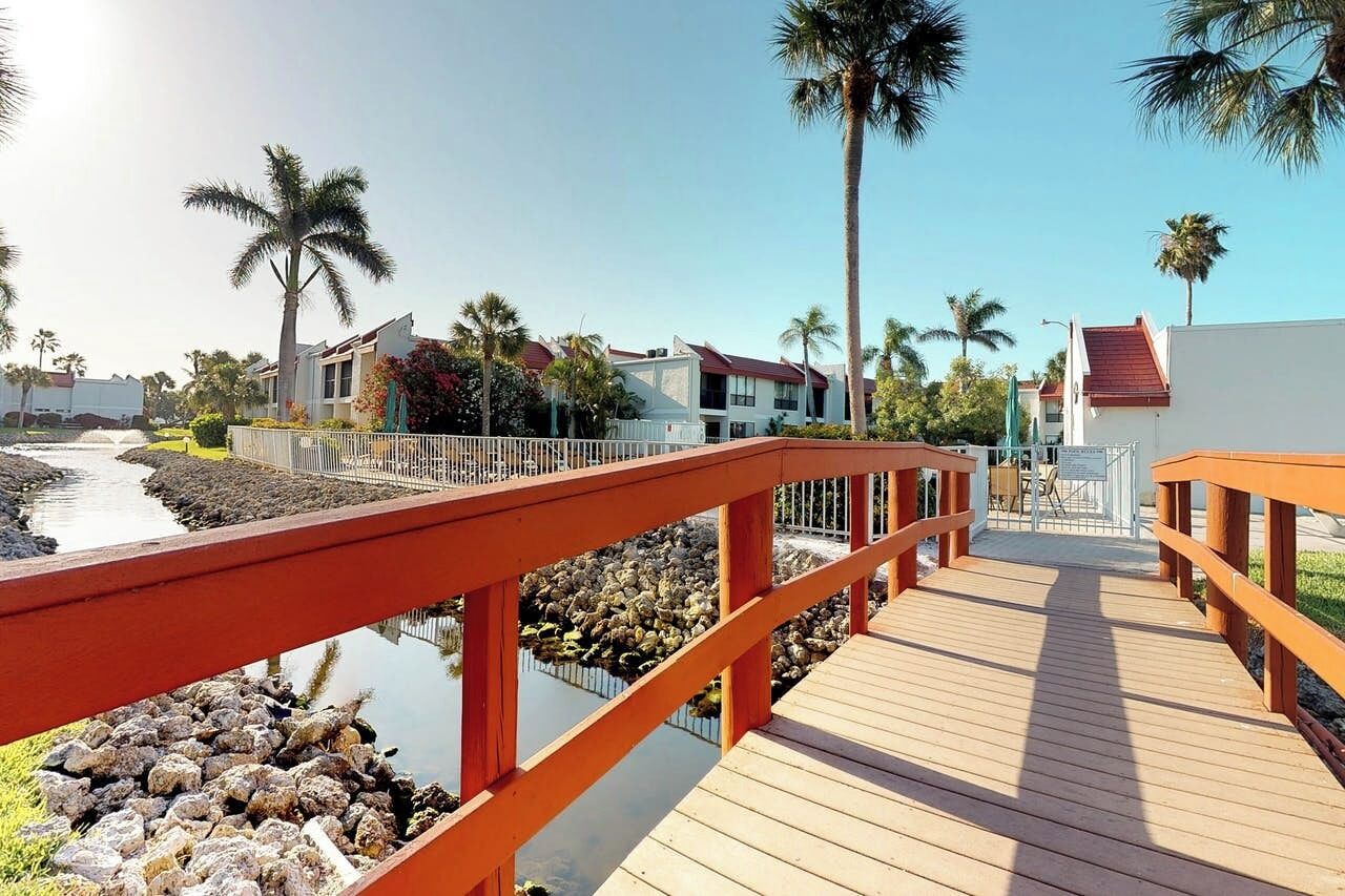 A wooden boardwalk leading to a building with a red roof, surrounded by palm trees and a calm water body.