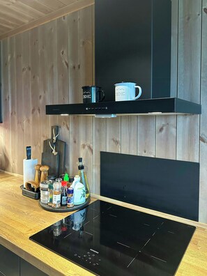 A kitchen with a black induction cooktop, wooden panel walls, a black shelf with two mugs, and various kitchen items on the counter. - Sunny retreat on Nesfjellet near alpine skiing (Buskerud)