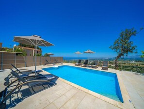 A swimming pool with lounge chairs and umbrellas, surrounded by a fence, with a view of the landscape. - Villa Candice With Private Pool (Rethymno)