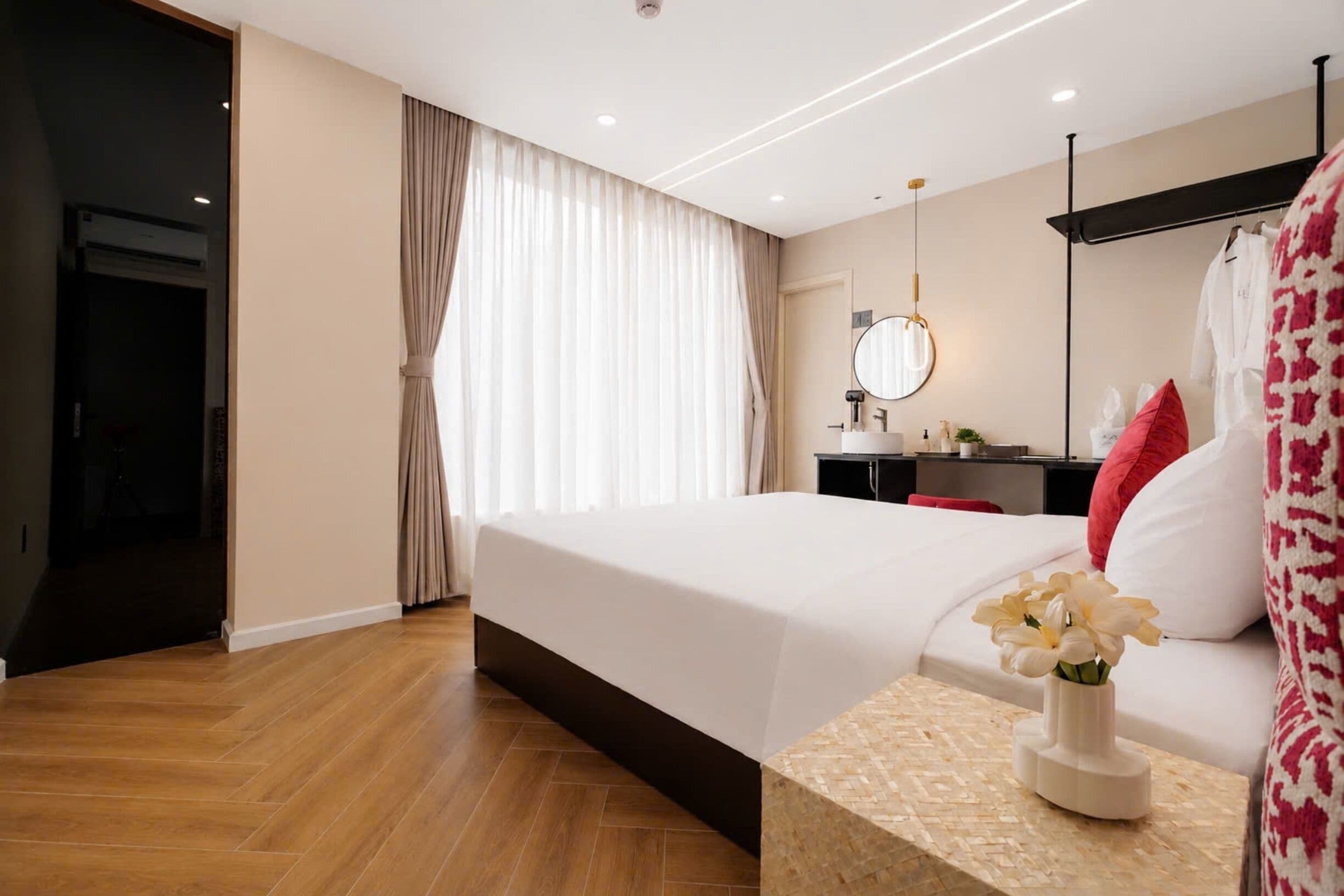 A modern hotel room with a large bed, a bedside table with a vase of flowers, a desk with a mirror, and a wardrobe.