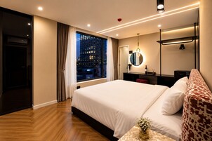 A hotel room with a large bed, a bedside table with a vase, a mirror, a wardrobe, and a view of the city at night.