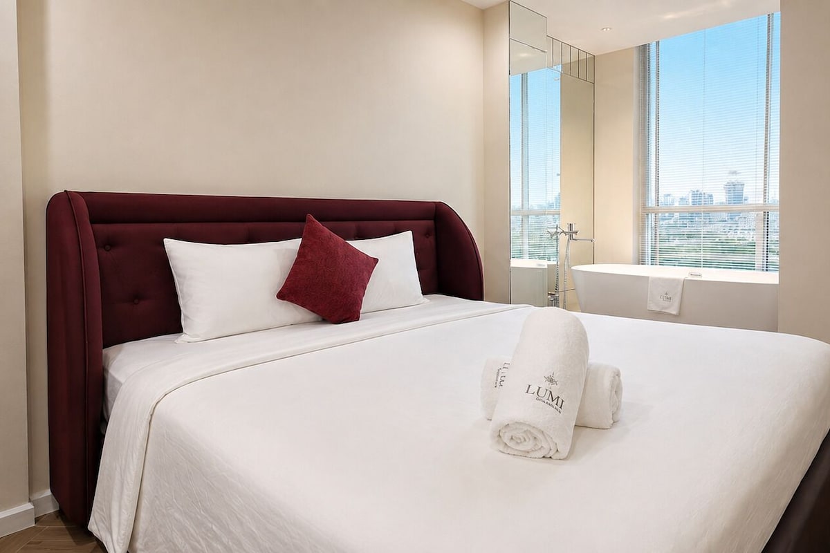 A hotel room with a bed, a red headboard, a window with a city view, and a towel with 'LUMI' branding.