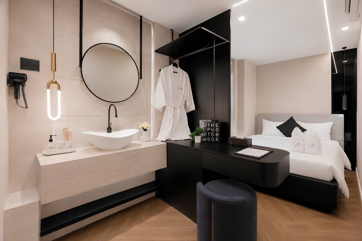 A modern hotel room with a large round mirror, a black vanity, a bed with white bedding, and a small seating area.