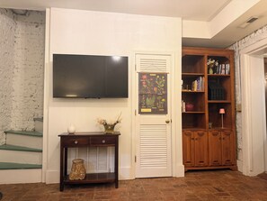 A room with a flat-screen TV mounted on the wall, a wooden bookshelf, a small table with decorative items, and a door with a botanical print.