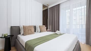Una habitación de hotel con cama, mesita de noche, lámpara y vistas a la ciudad a través de amplios ventanales.