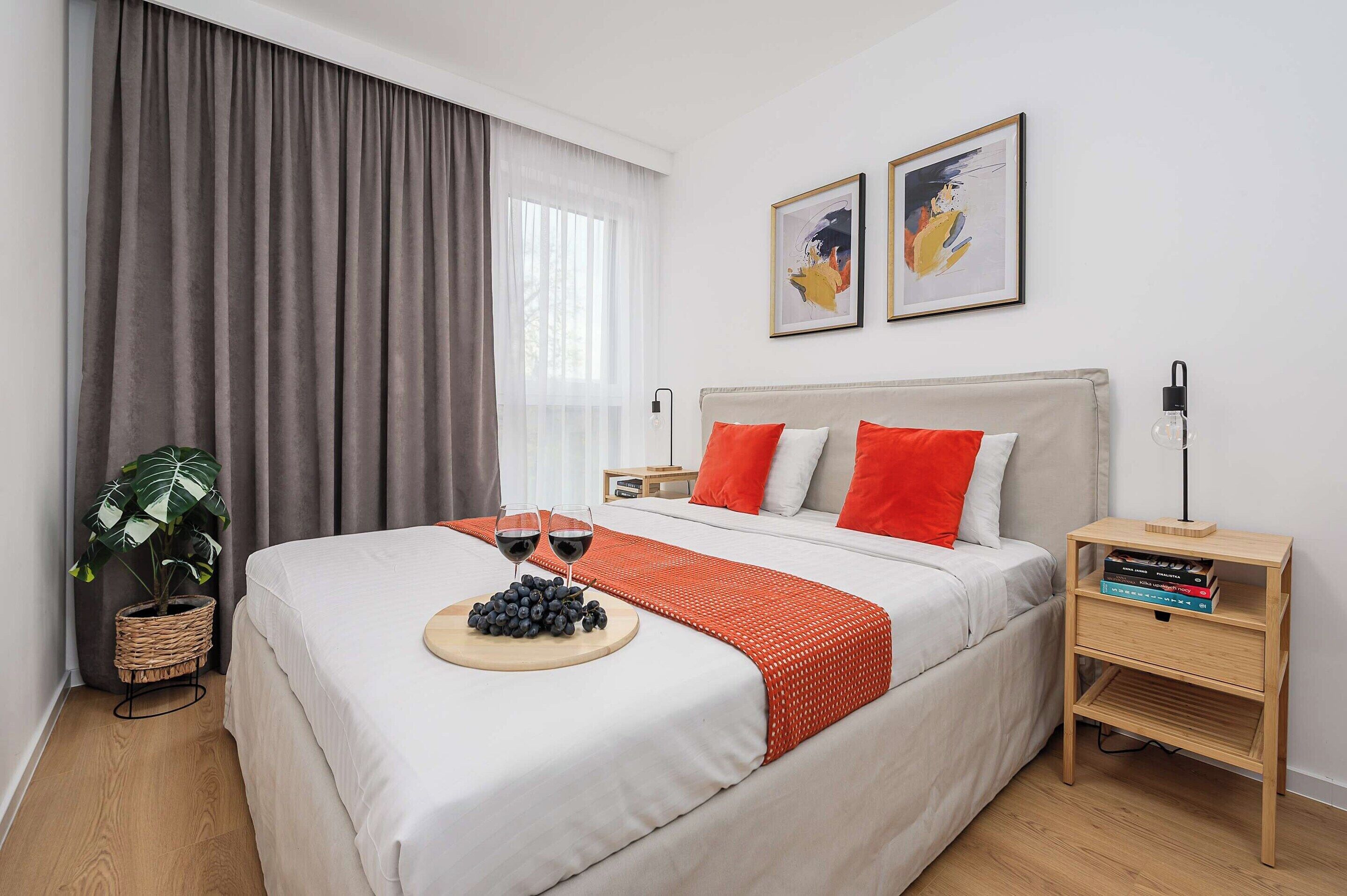 Une chambre d’hôtel avec un lit, une table de chevet avec des livres, une plante et une fenêtre avec des rideaux.
