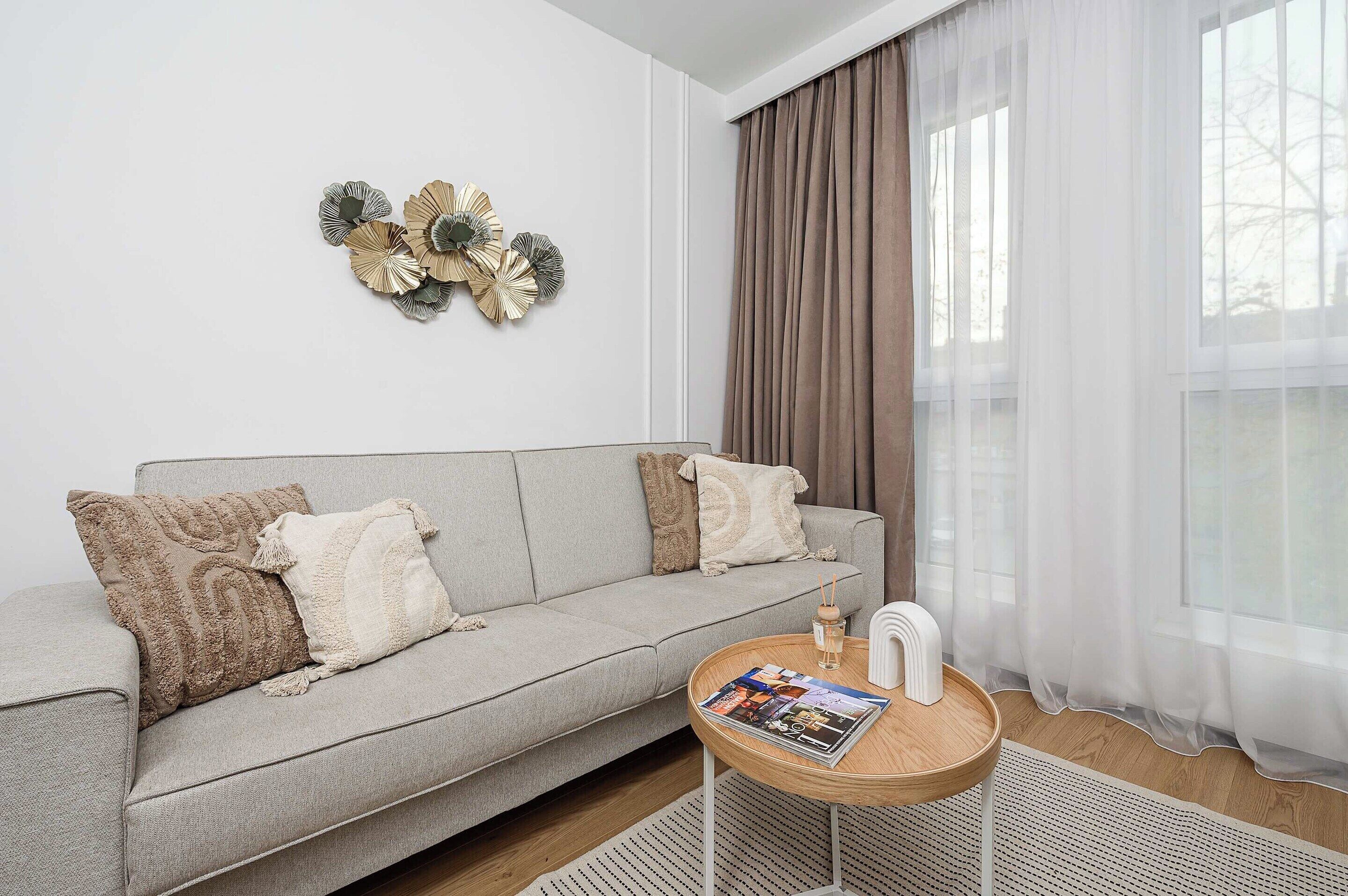 A modern living room with a grey sofa, decorative wall art, a round wooden coffee table with magazines, and sheer curtains.