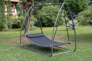 Een metalen schommelstoel met een zwarte zitting en rugleuning, geplaatst op gras met bomen en een gebouw op de achtergrond.