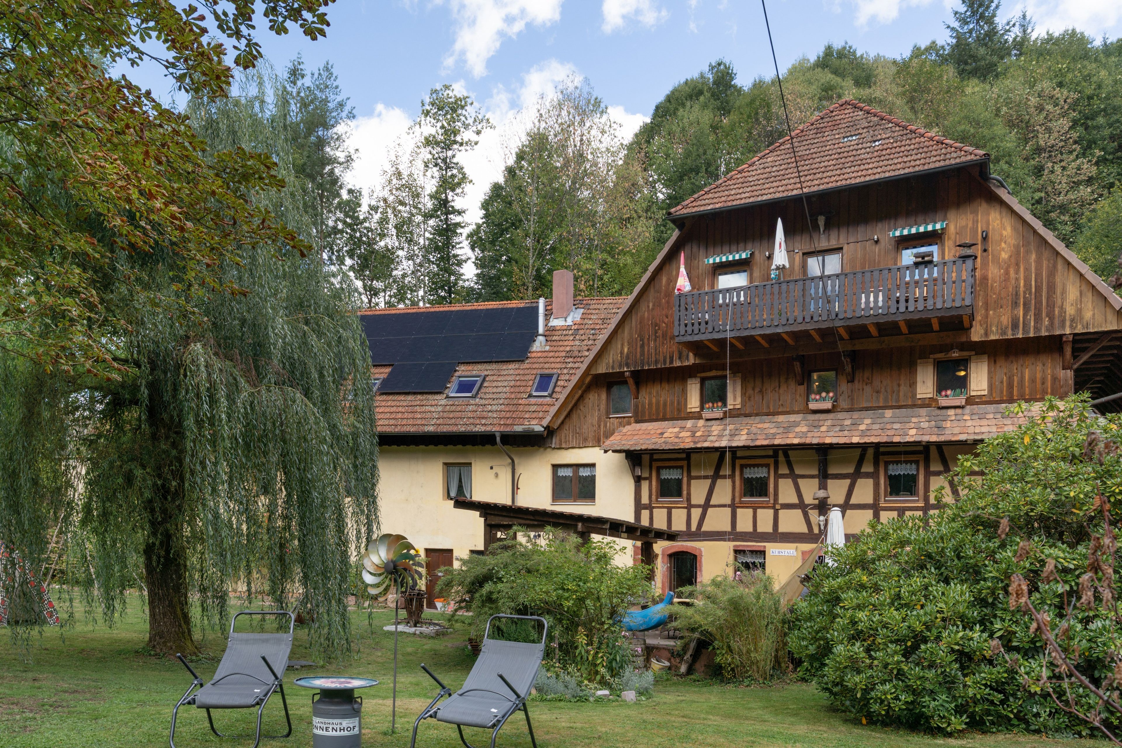 A traditional wooden chalet with a balcony, surrounded by lush greenery and a clear sky.