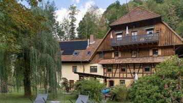 A traditional wooden chalet with a balcony, surrounded by lush greenery and a clear sky.