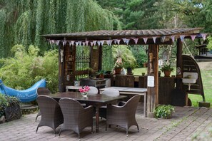 A rustic outdoor seating area with a wooden table and wicker chairs, surrounded by greenery and a blue banana-shaped decoration.