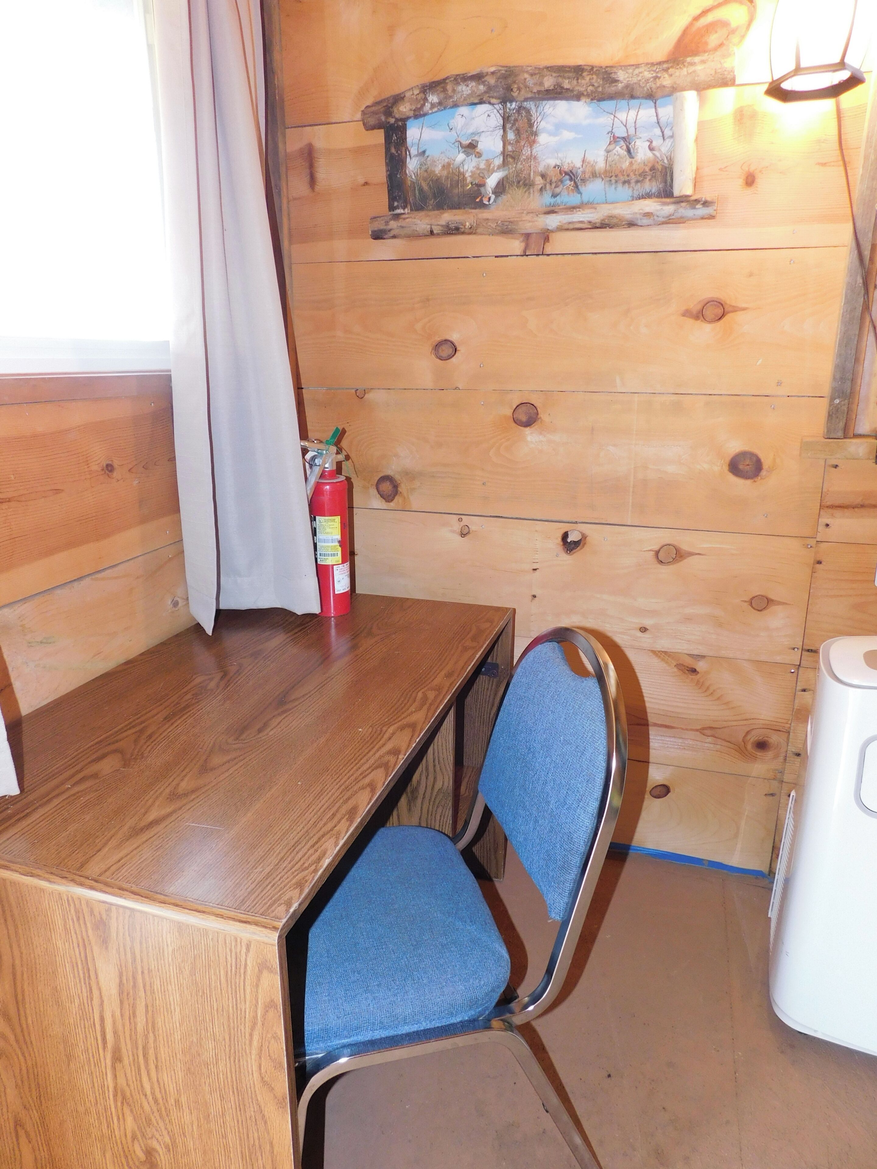 A wooden cabin interior with a dining table, a blue chair, a fire extinguisher, and a rustic wall painting.