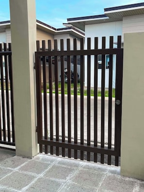 A gated entrance with a metal fence, a paved driveway, and a building with a flat roof in the background.
